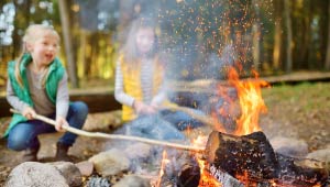 A picture with a focused view of a campfire, with two kids (blurred) in the background poking the fire with sticks.